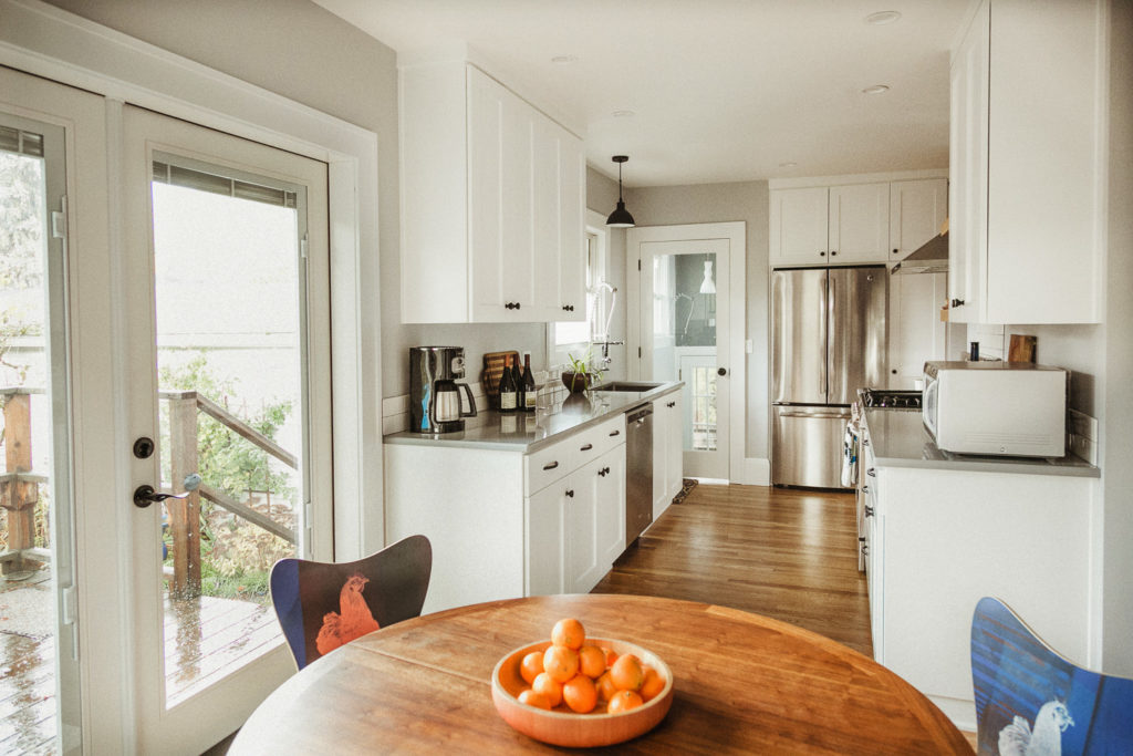Hardwood floors, white cabinetry, light grey countertops, french doors to back deck. Natural wood round table with funky chairs with chicken images on them. Stainless steel appliances.