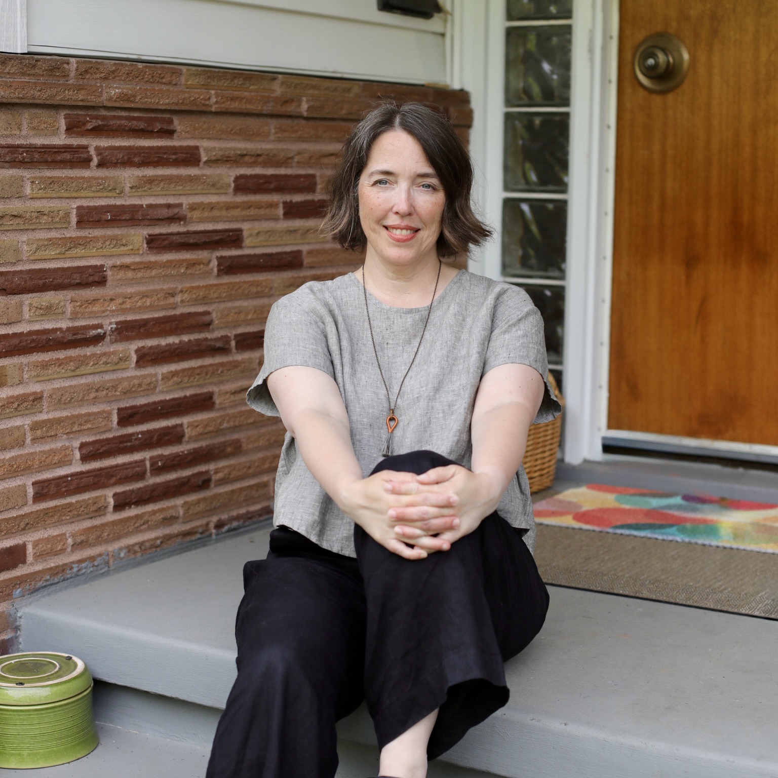 Amanda Erickson bio pic, seating on front steps in front of brick wall and wood door, 1950s home.