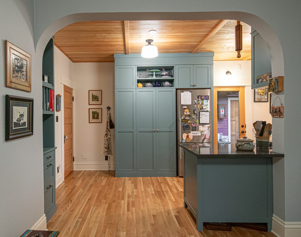 Wood floors and ceiling, full height pantry and refrigerator surround, teal cabinetry in a shaker style.