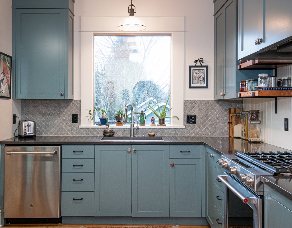 Teal cabinetry, oversized window, mid-tone grey scalloped tile. Undermount sink with pull-down faucet.