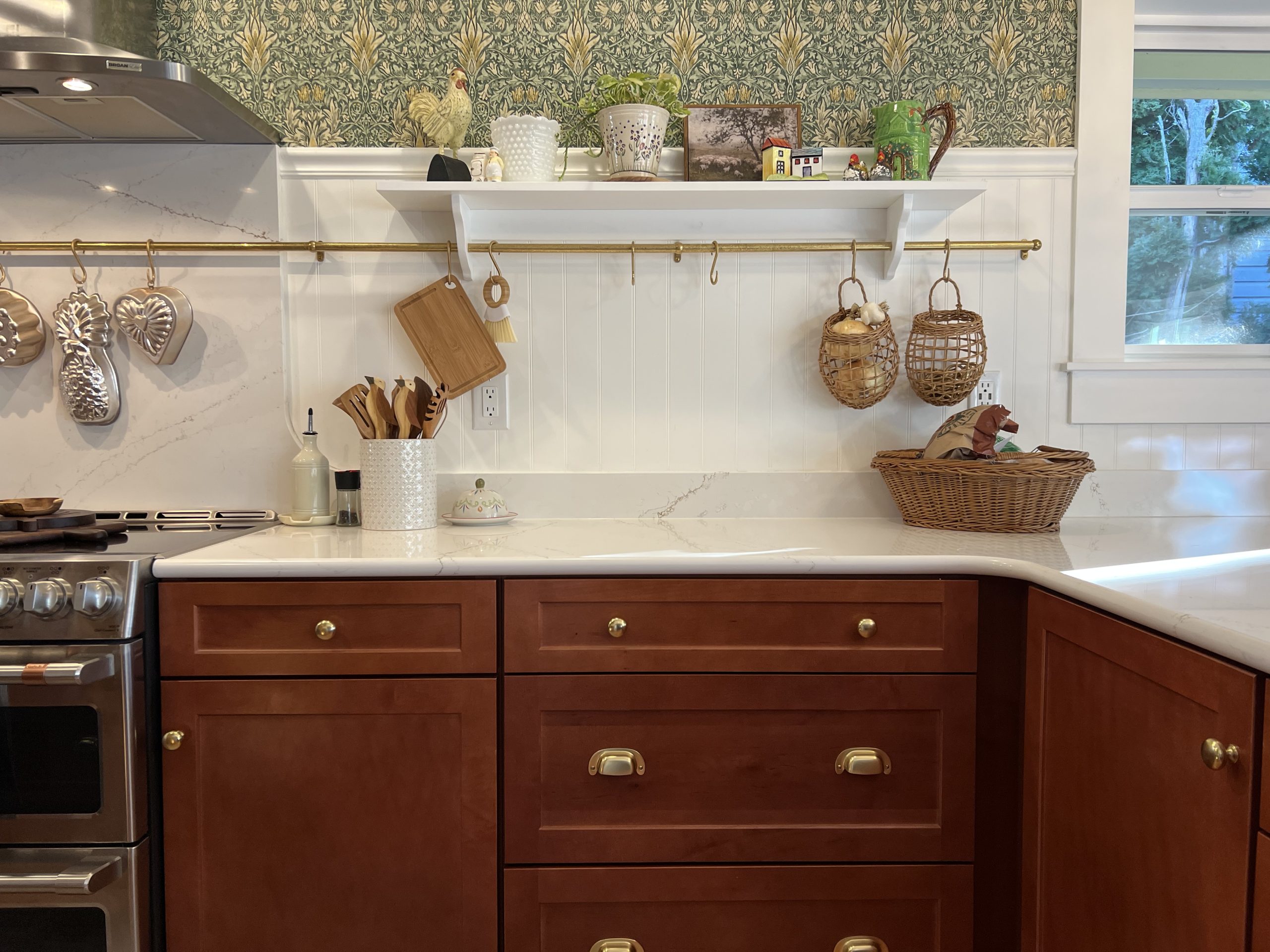 Kichen countertop detail with a brass hanging rail, decorative shelf, white wainscoting and William Morris style wallpaper above.