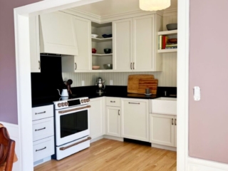 Lavender walls and a white painted trim archway frame the kitchen. White painted cabinetry. Black countertops and backsplash. Farmhouse Sink.