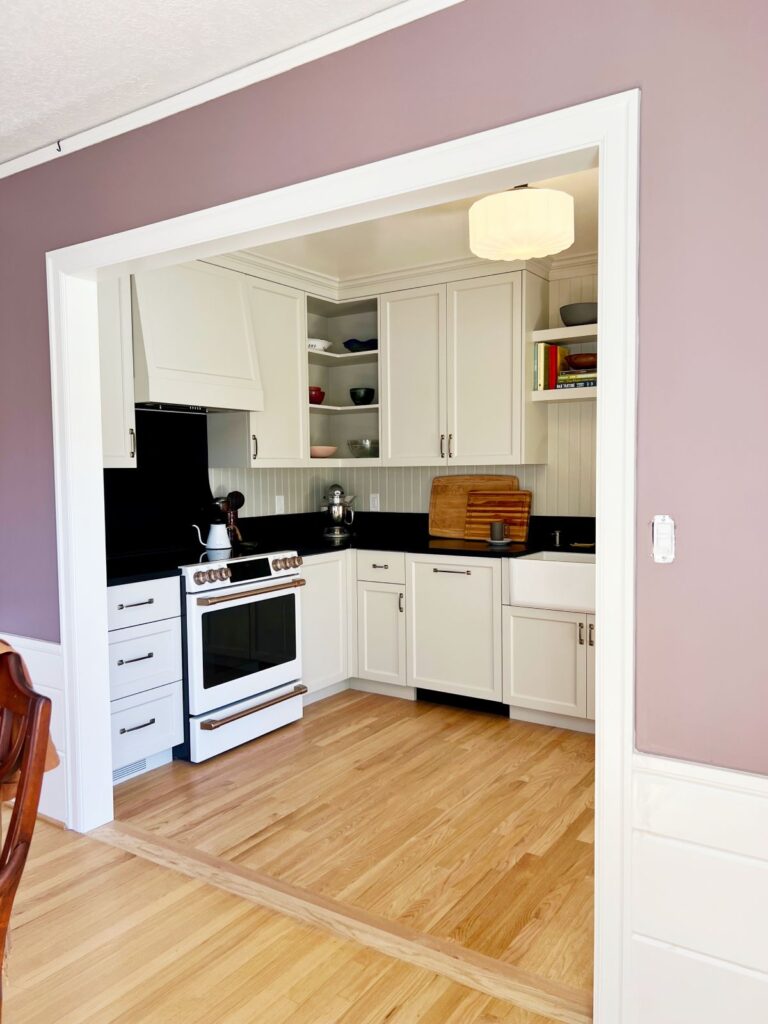 Lavender walls and a white painted trim archway frame the kitchen. White painted cabinetry. Black countertops and backsplash. Farmhouse Sink.