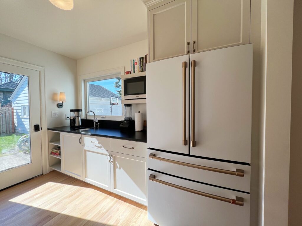 Light-filled room, cream cabinetry. White refrigerator with copper accents. Black countertop and backsplash.