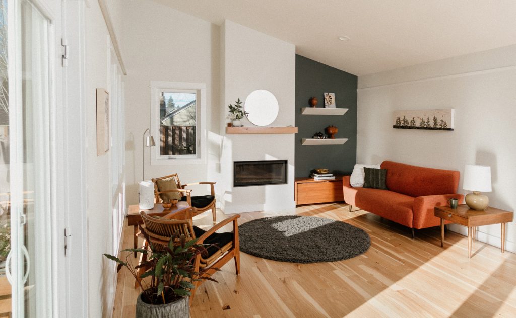 Light wood floors, circular dark grey area rug, dark grey accent wall, mid-mod furniture, electric hearth with a floating wood mantel above and small circle mirror