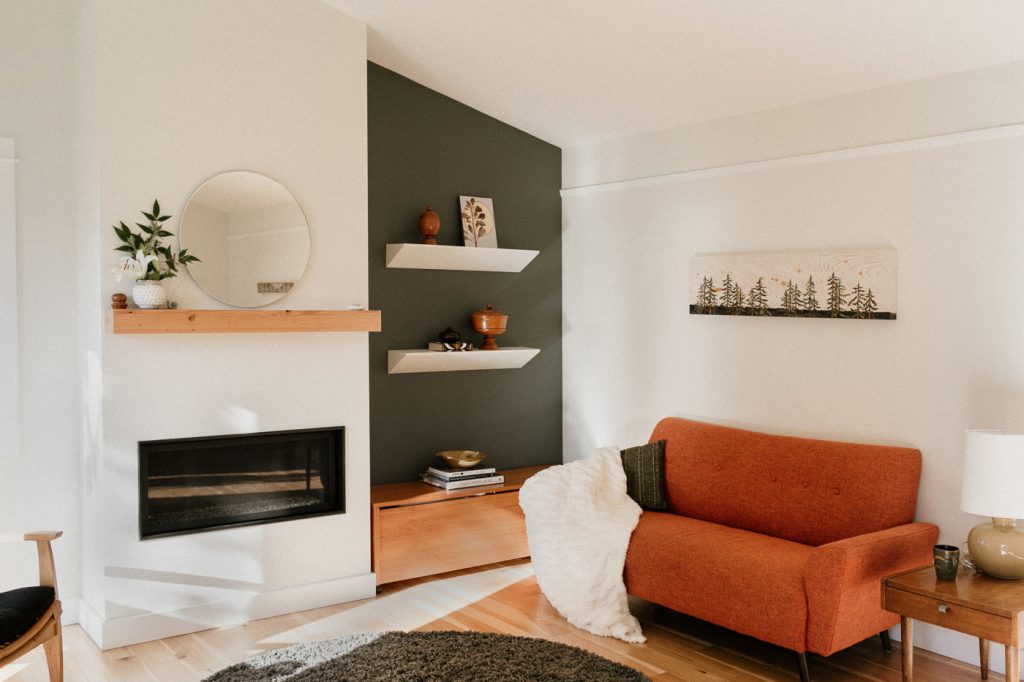 Light wood floors, white walls, with dark grey accent wall. Mid-mod furniture. Floating shelves with artwork and charming objects.
